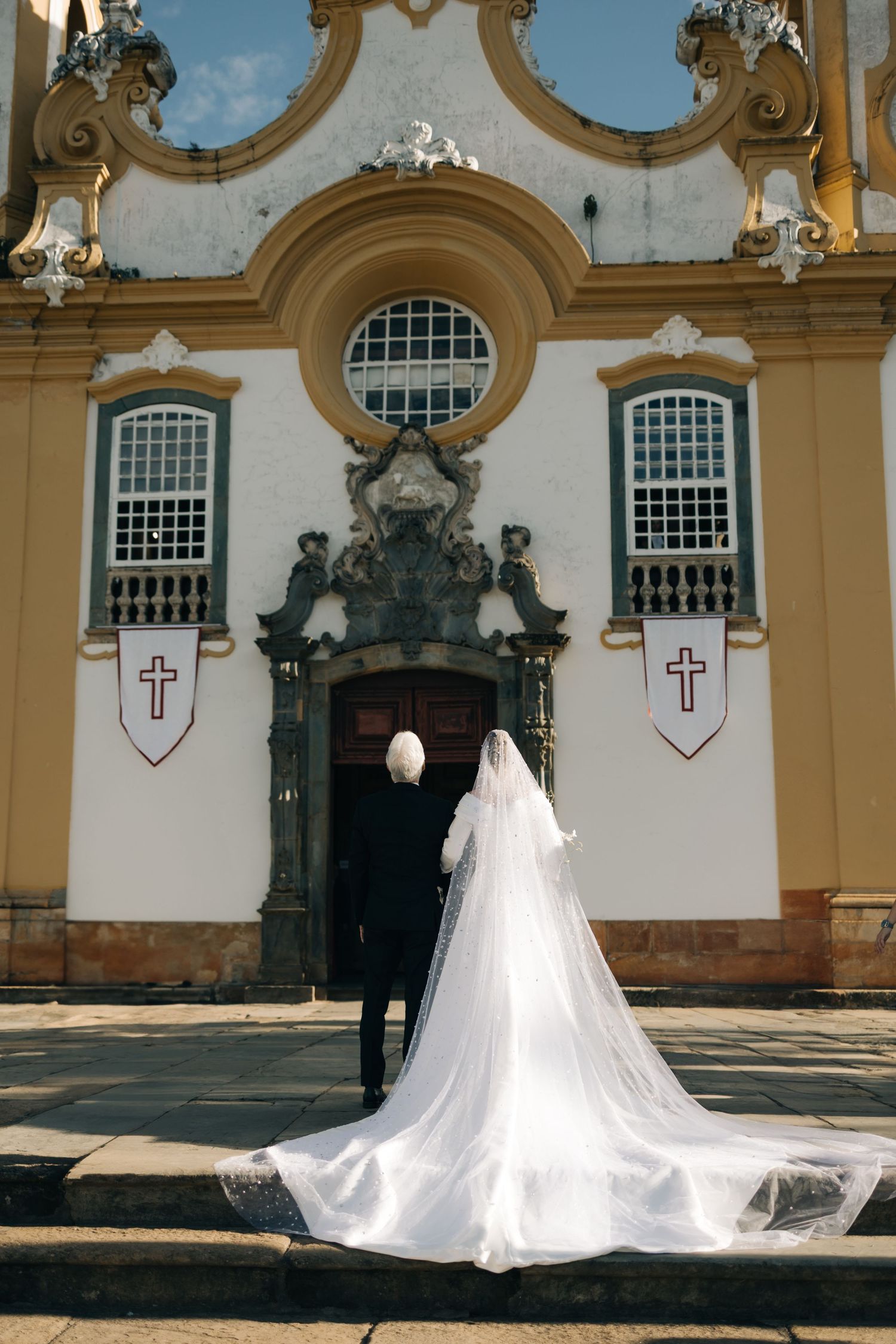 A bride in a flowing white gown with long veil stands at the entrance of a baroque church.