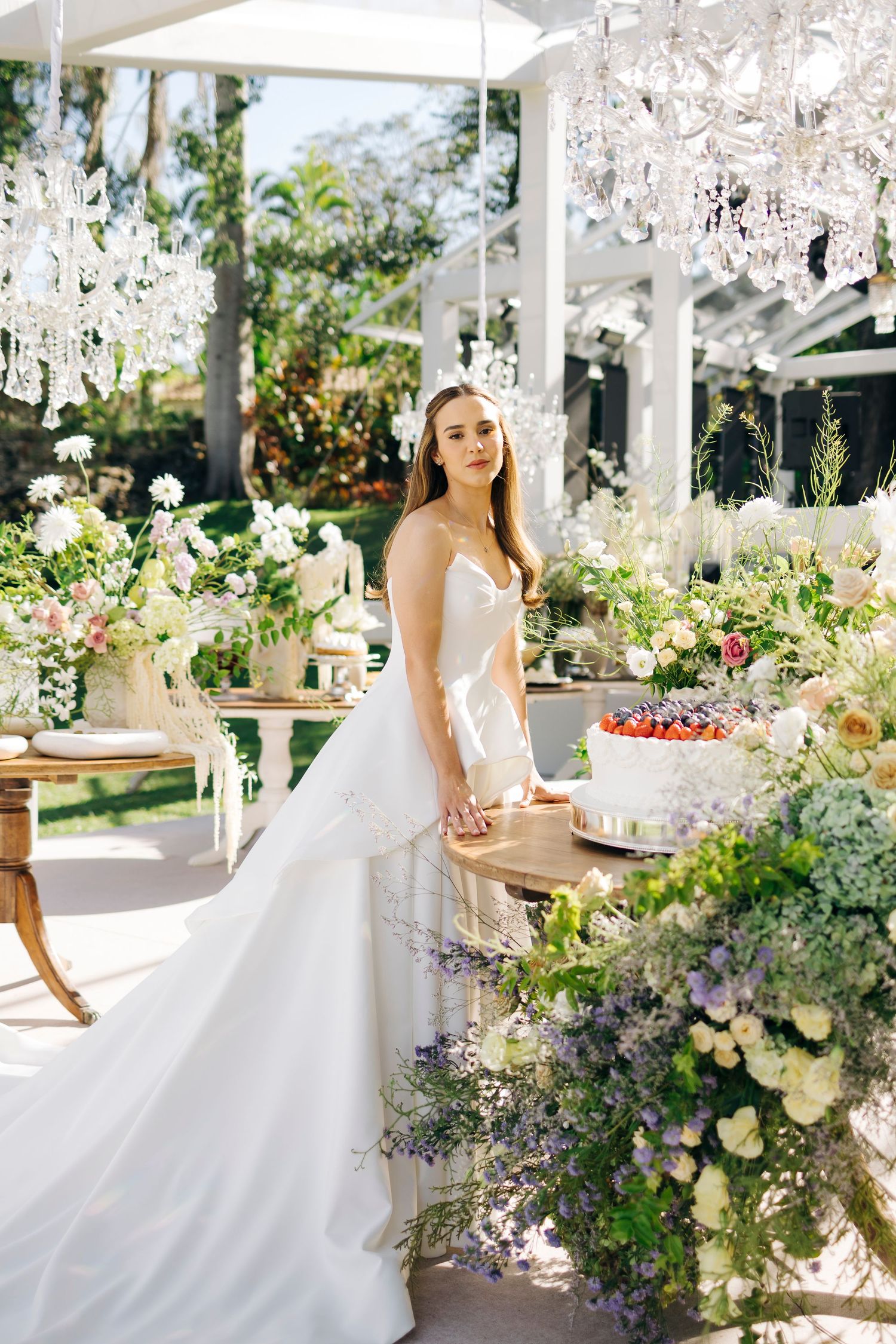 Bride in elegant white gown stands by floral-adorned wedding table with cake, crystal chandeliers, and lush garden backdrop.