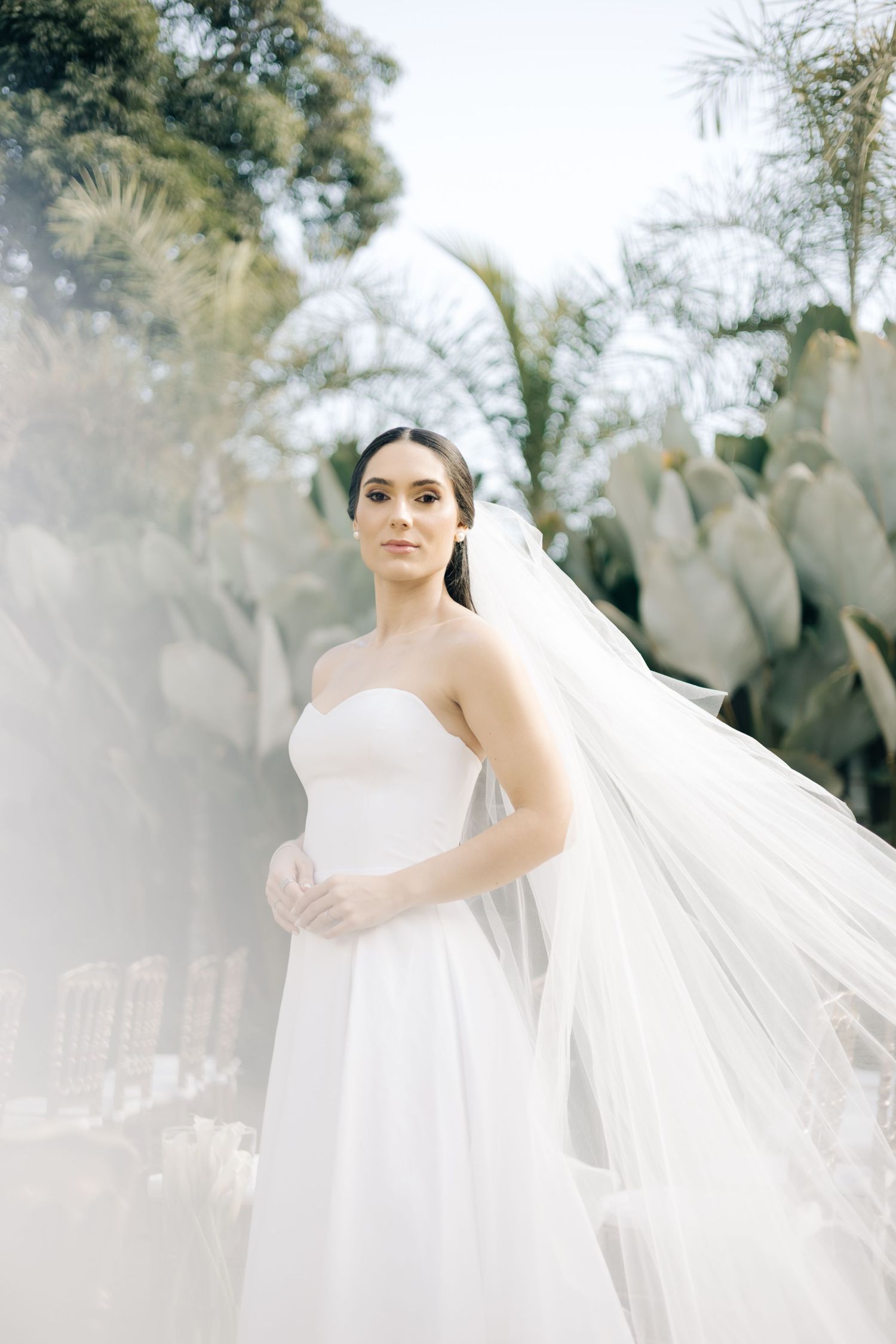 A bride in a flowing white dress and long veil stands elegantly in a garden setting with soft, dreamy lighting.