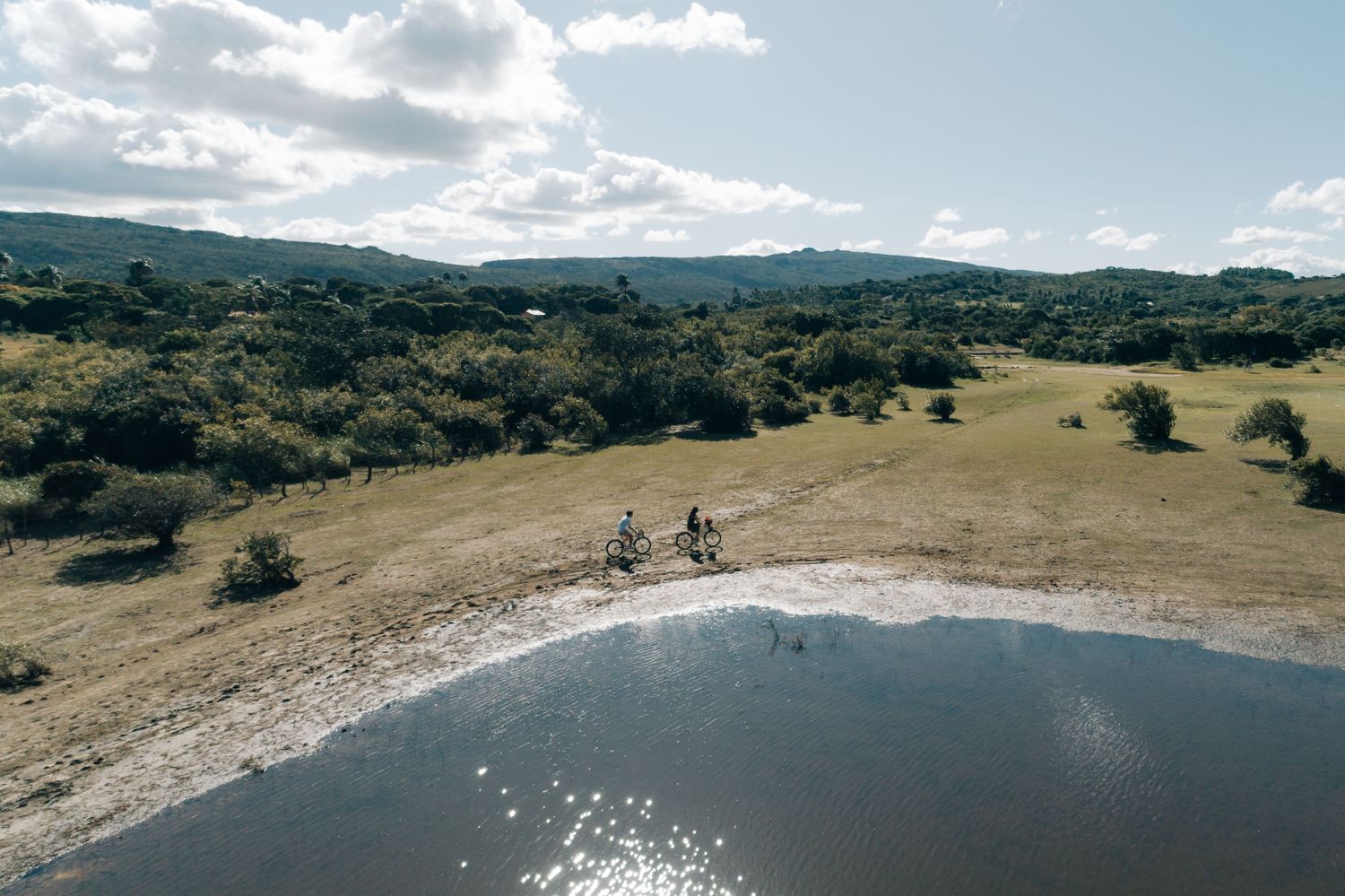 Two cyclists ride along a scenic trail overlooking a glittering lake surrounded by rolling hills on a sunny day.