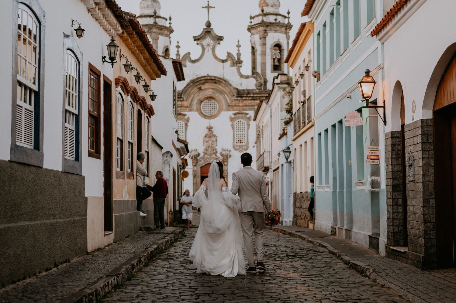 Wedding couple walking on a historic cobblestone street towards a baroque church facade.
