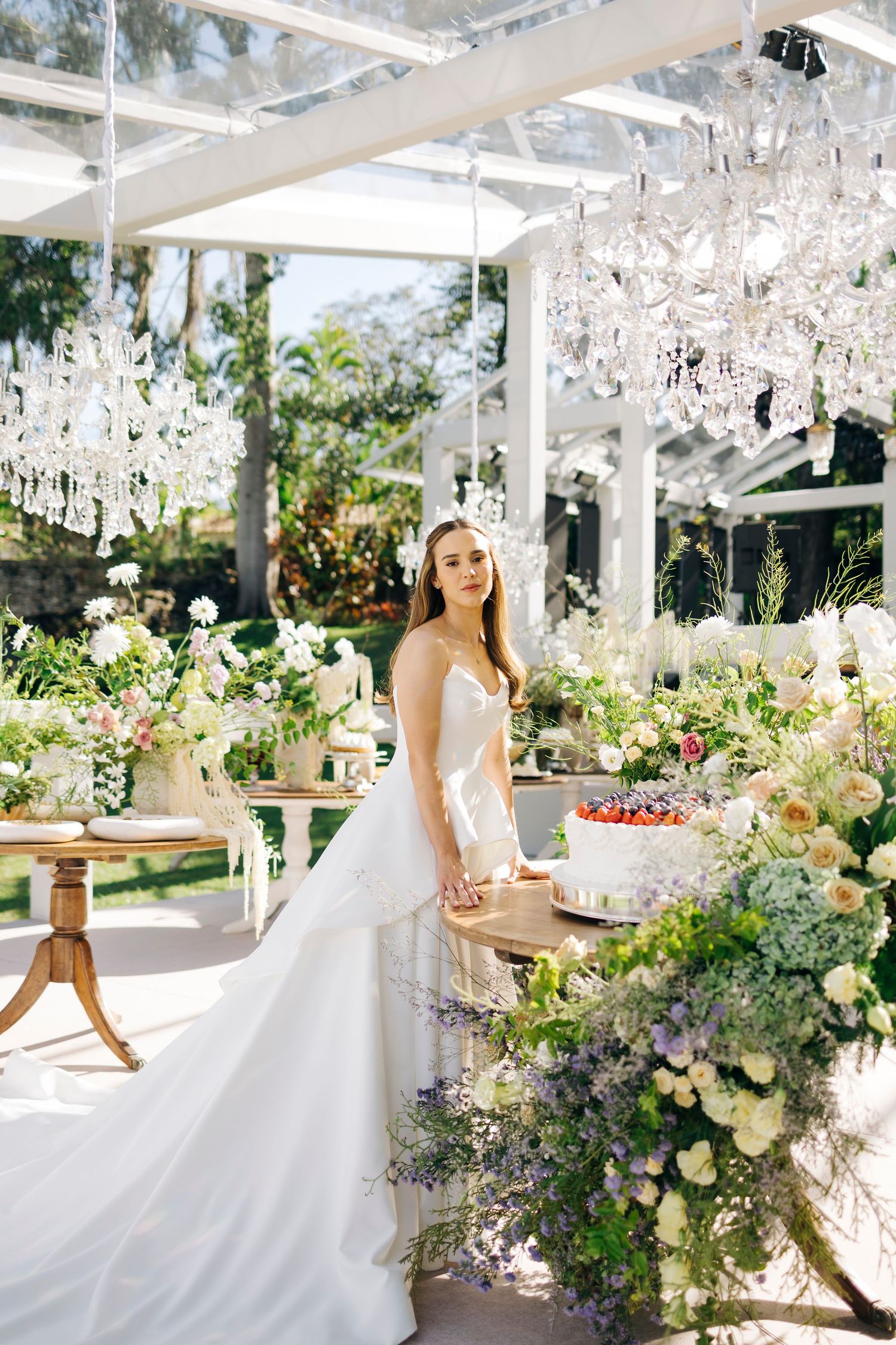 Bride in white gown stands beside floral wedding cake display under crystal chandeliers in elegant outdoor venue.