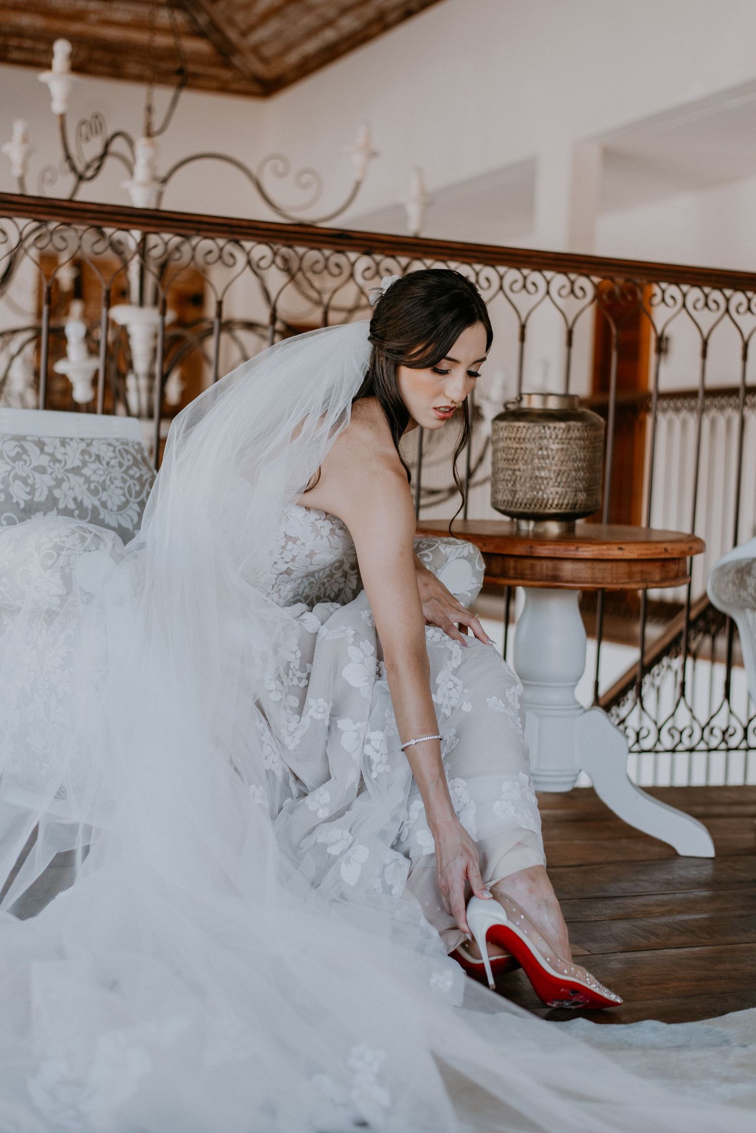 Bride in white wedding dress adjusting her designer shoes with red soles while seated in ornate room.