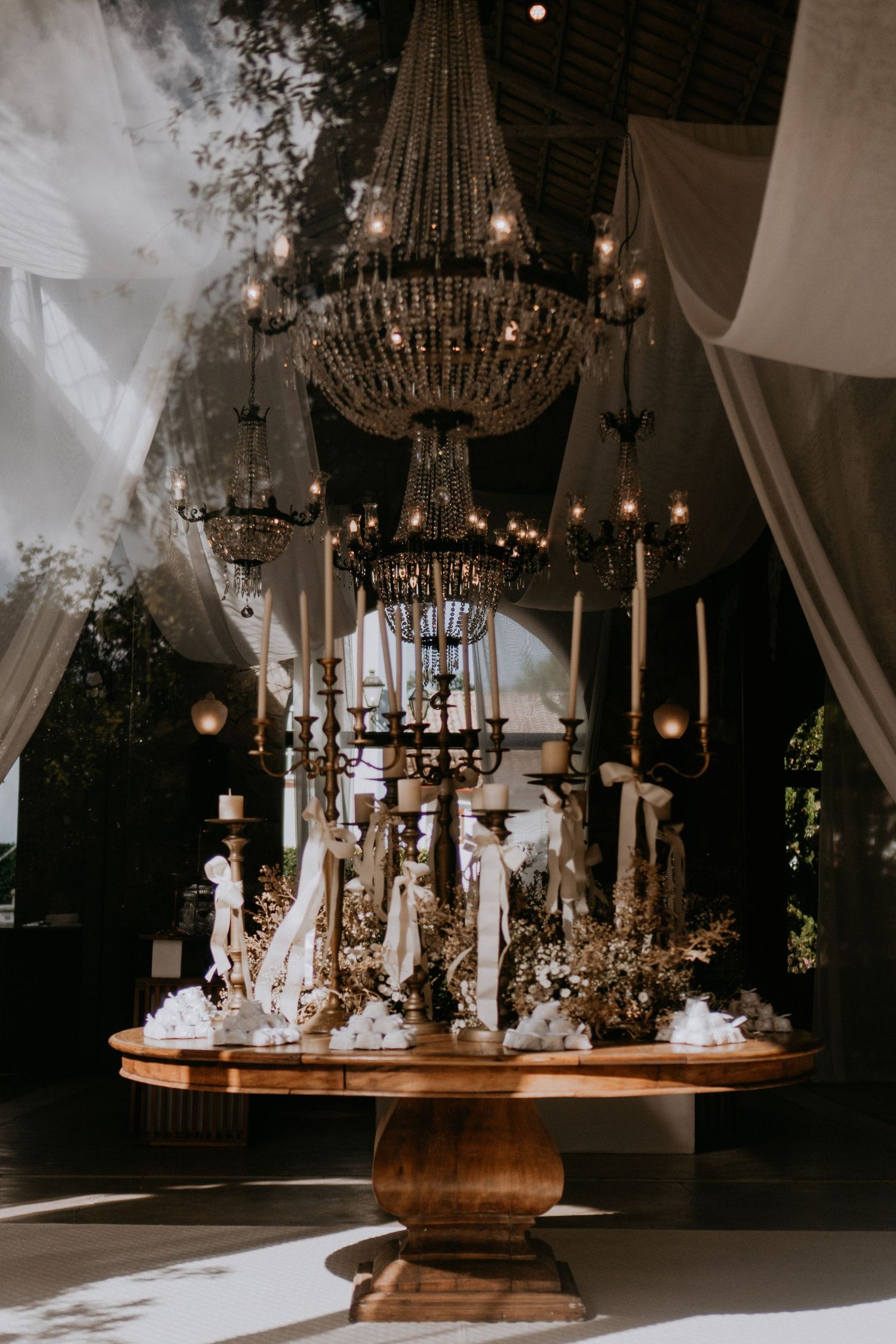 Ornate candle display on wooden pedestal table under crystal chandeliers and draped fabric.