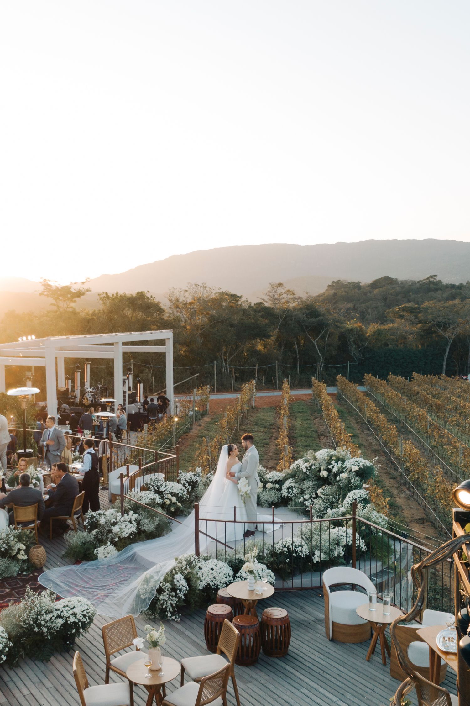 Couple shares a romantic kiss at sunset vineyard wedding on a wooden deck adorned with white floral arrangements.