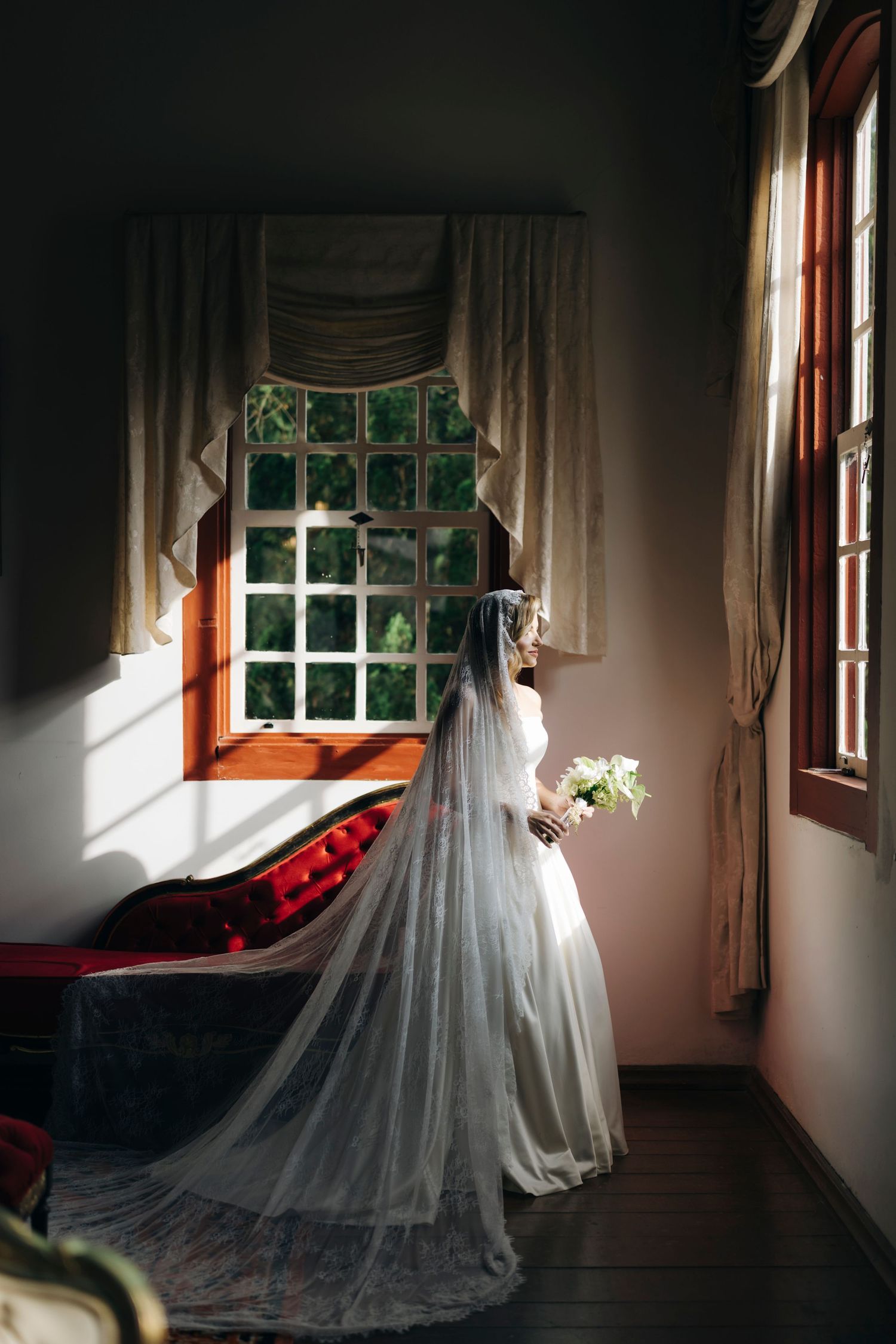 Bride in white wedding gown and veil holds bouquet while gazing out a sunlit window in an elegant room.