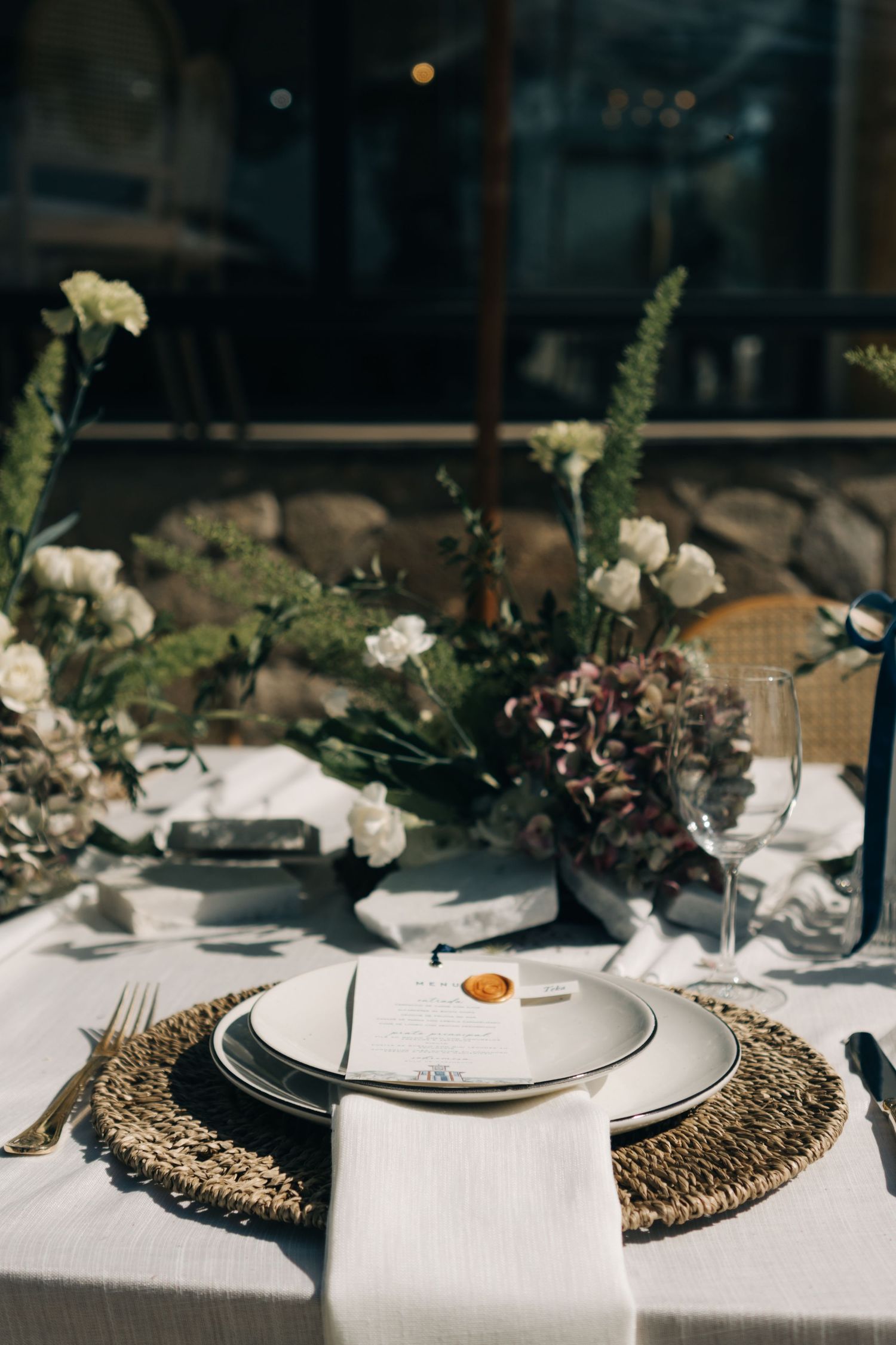 Elegant table setting with white plates on rattan placemats and floral centerpiece featuring white flowers and greenery.