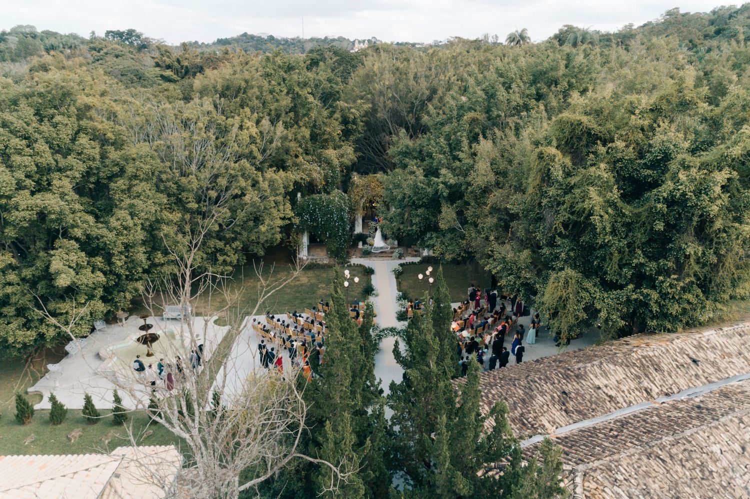 Aerial view of an outdoor wedding ceremony in a wooded area with guests gathered around a stone fountain.