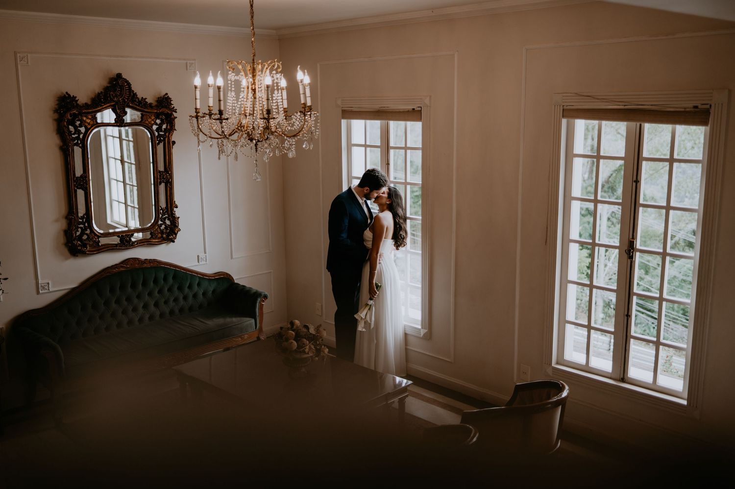 Two figures share an intimate moment in elegant room with vintage chandelier, ornate mirror and French doors.