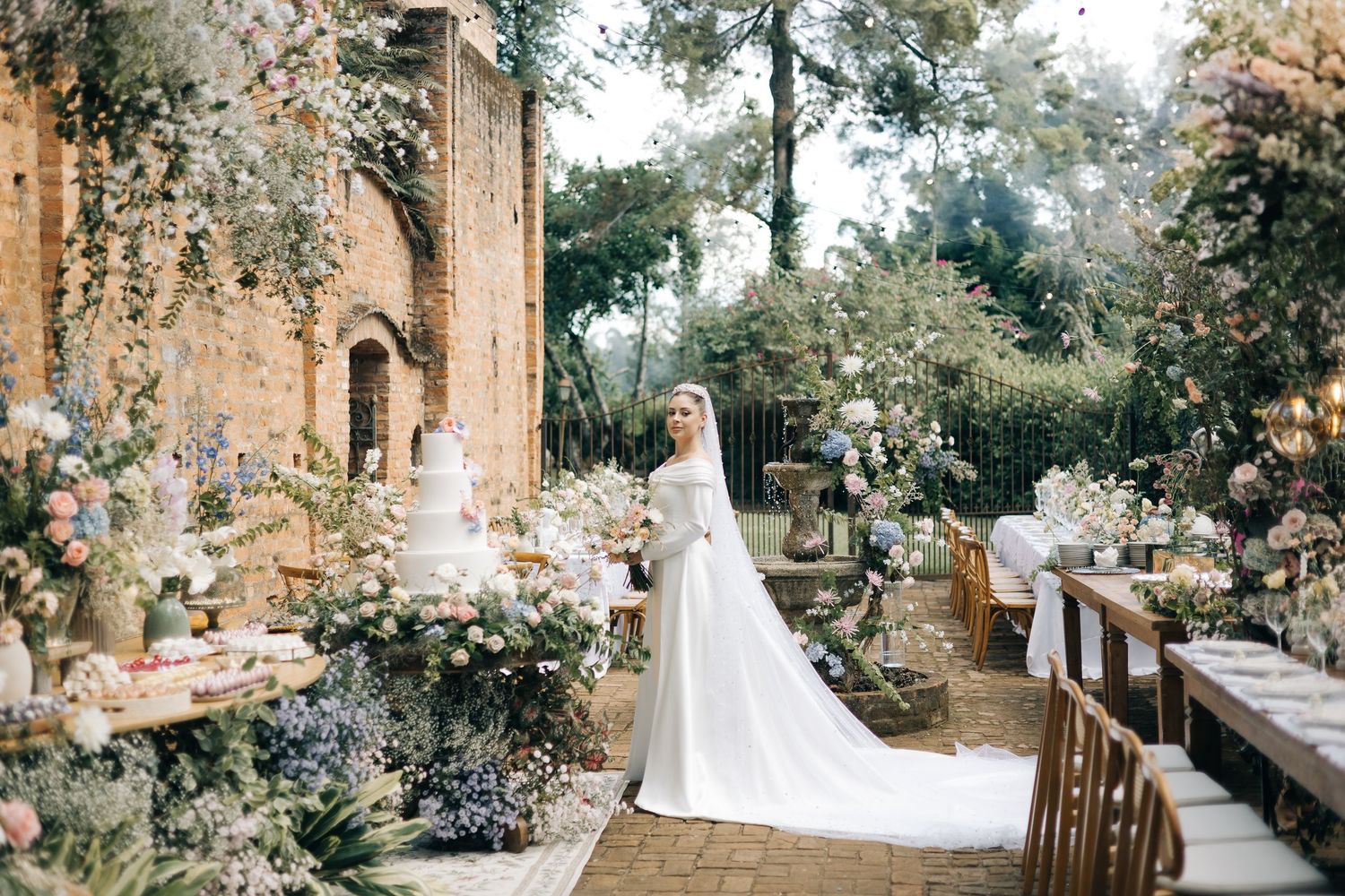 Bride in white gown with long train stands among lush floral arrangements at an elegant outdoor wedding venue.