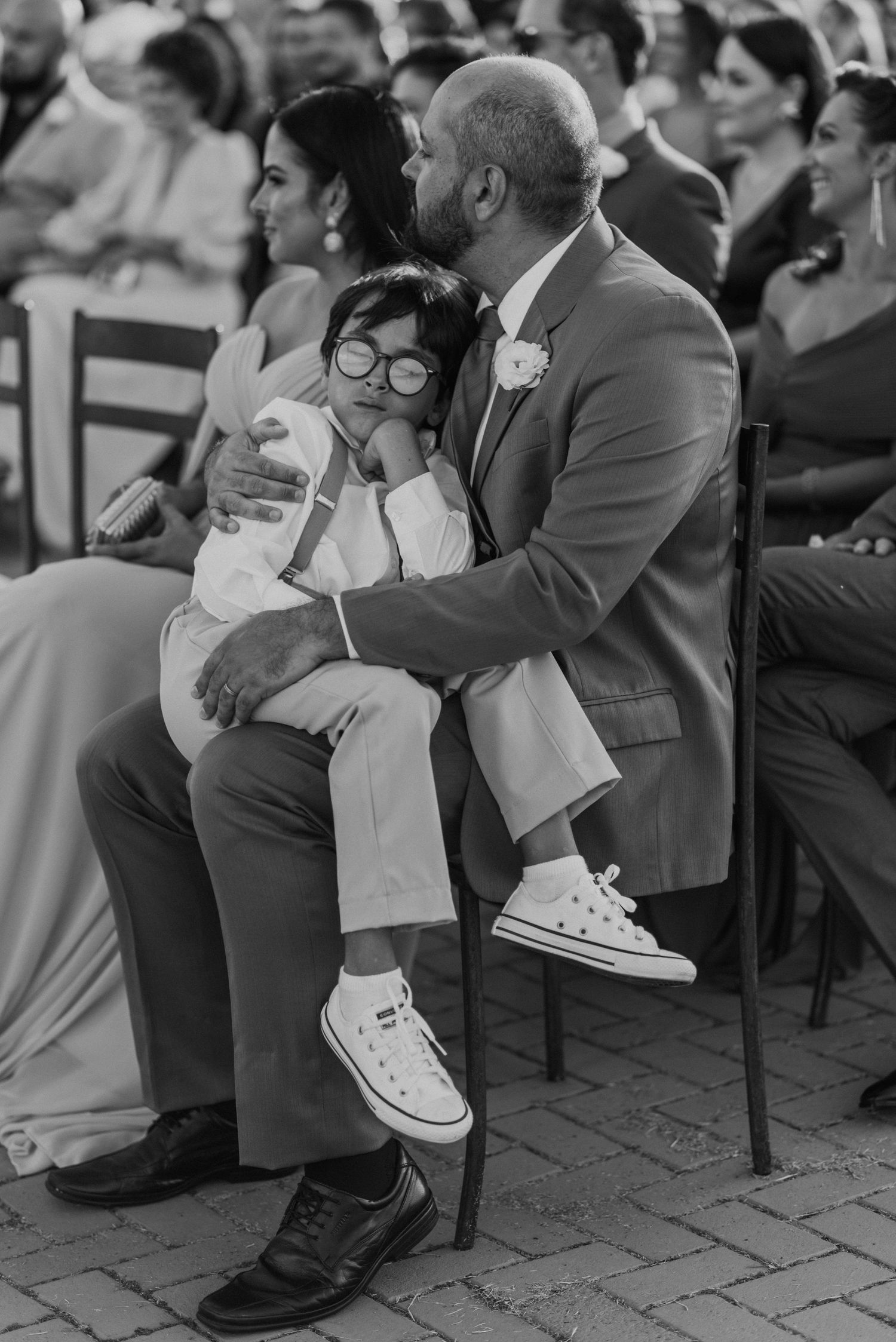Wedding guest in grey suit holds young child wearing white sneakers during ceremony.