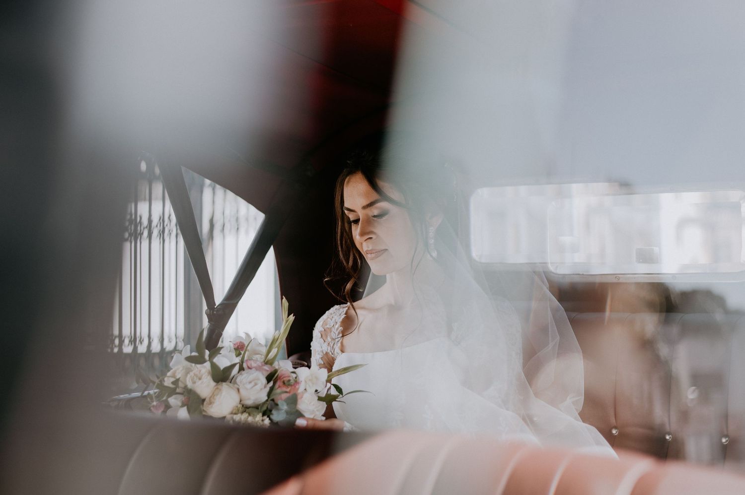 Bride with flowing veil and floral bouquet in ethereal wedding photography shot through glass.