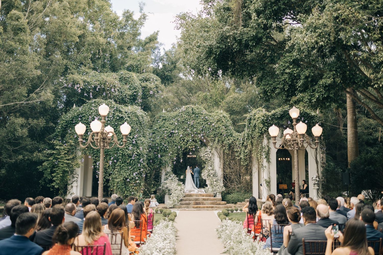 Garden wedding ceremony with guests seated beneath floral arches and decorative light posts.