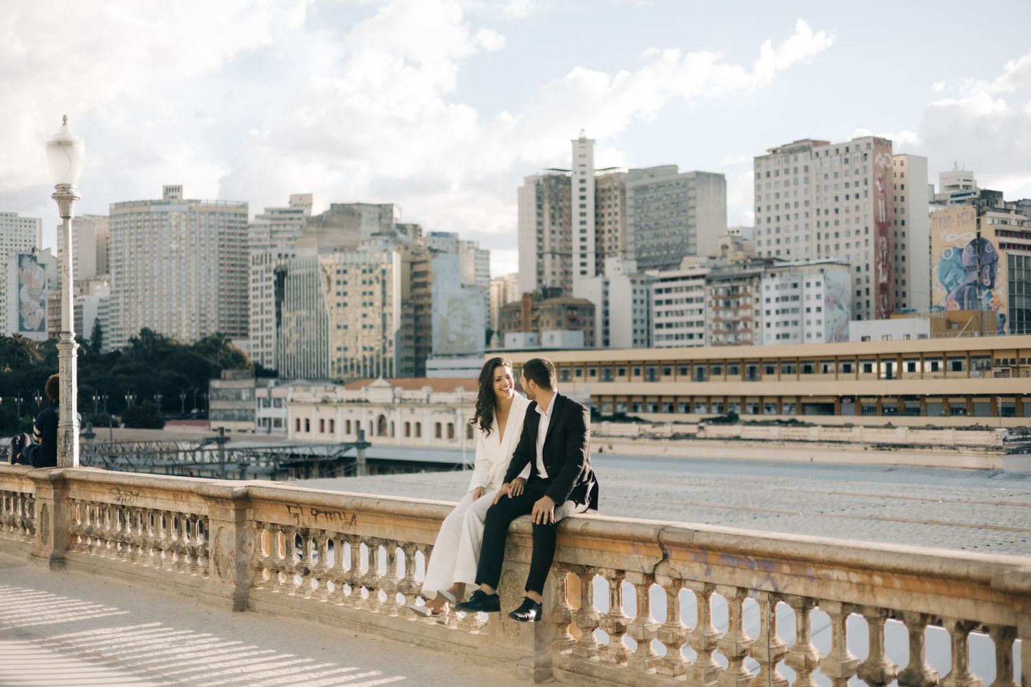 Person sits on ornate stone bridge railing with modern city buildings and cloudy skyline of Sao Paulo in background.