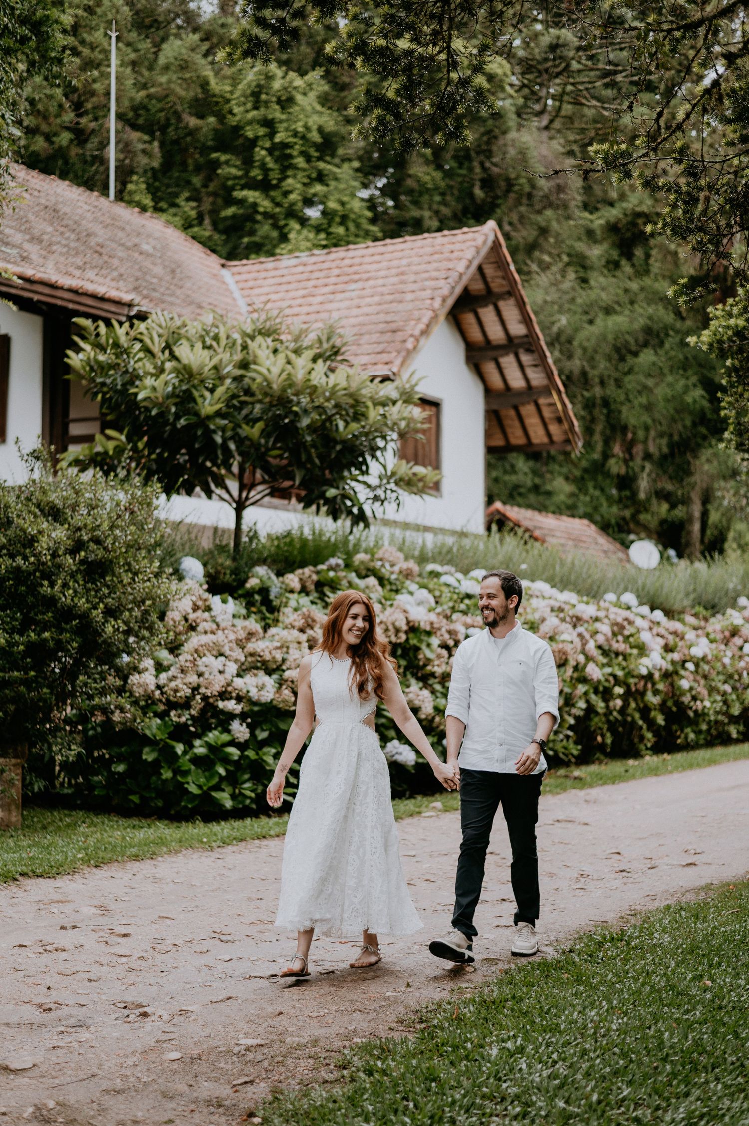 Two people walking on a path surrounded by lush garden flowers and a white cottage house in a rustic countryside setting.