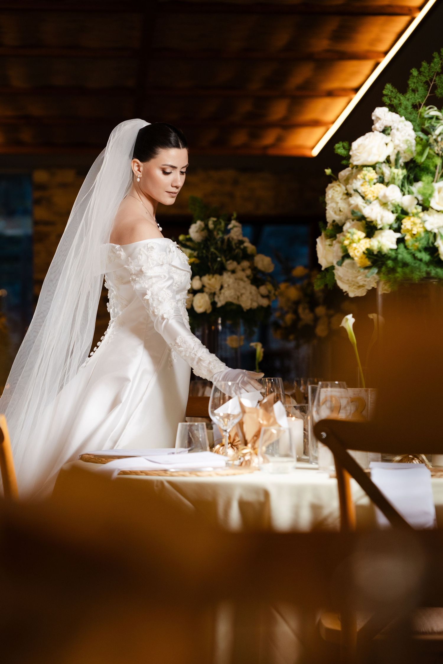 Elegant bride in off-shoulder lace gown with veil arranges reception table with flowers.
