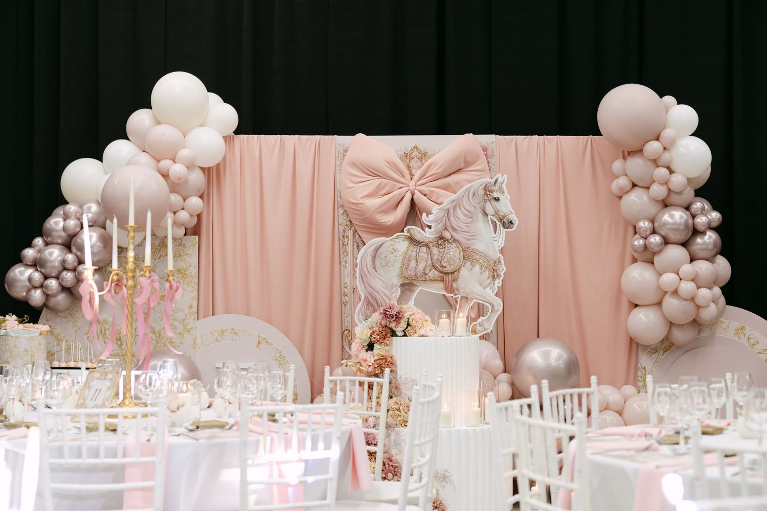Elegant pink and white christening party decor with carousel horse, balloons, and decorated tables.