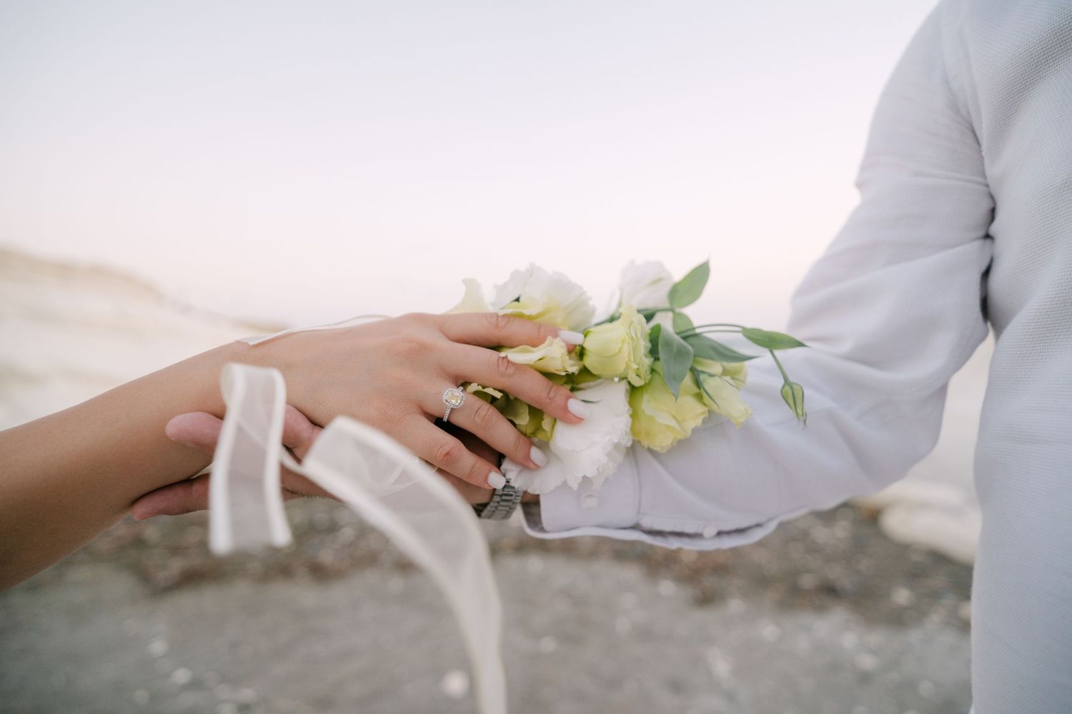 Close-up of couple holding white floral bouquet with ribbon on a sunlit rocky beach.