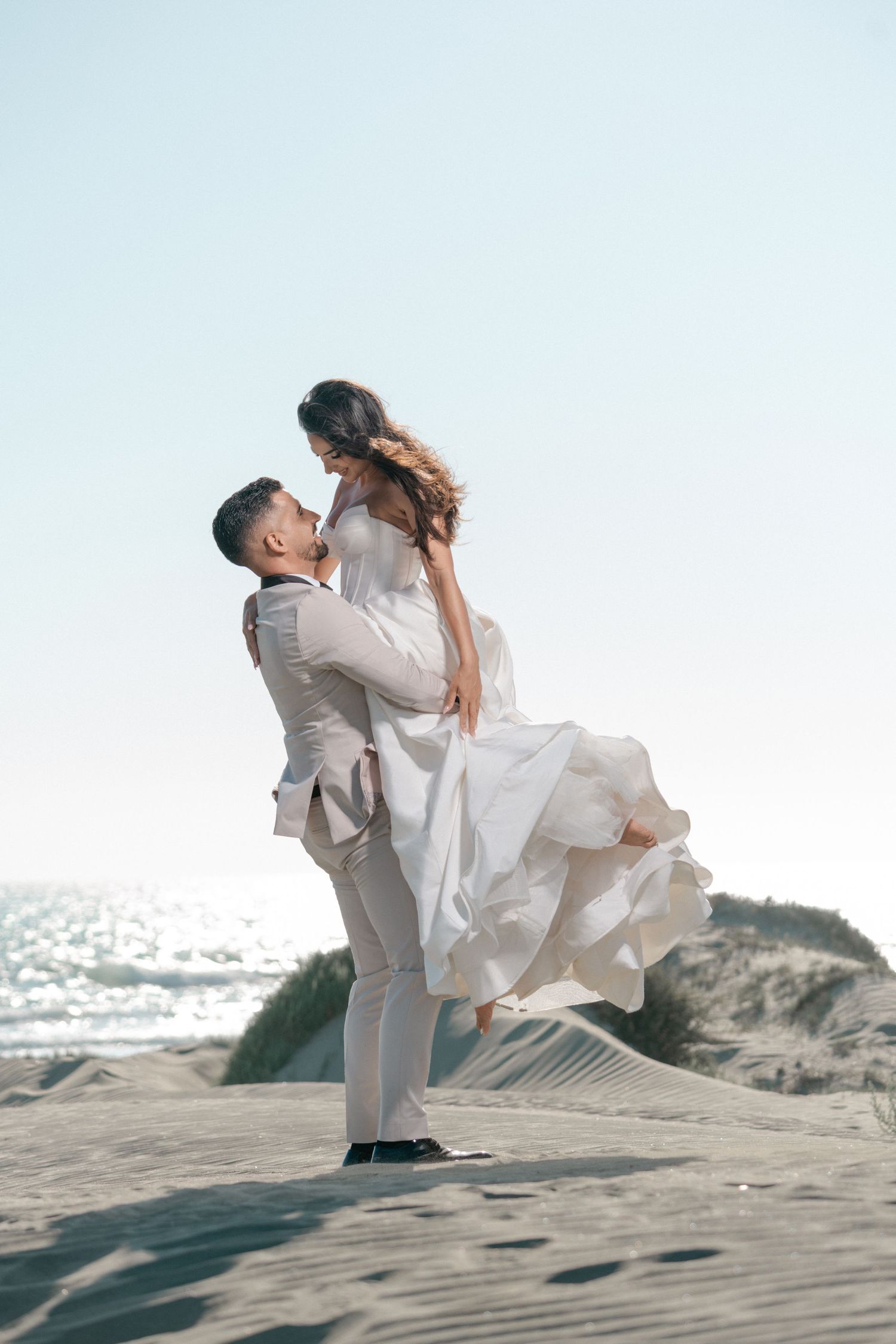 Groom lifts bride in a white dress on a sandy beach, both dressed in white with the ocean backdrop.