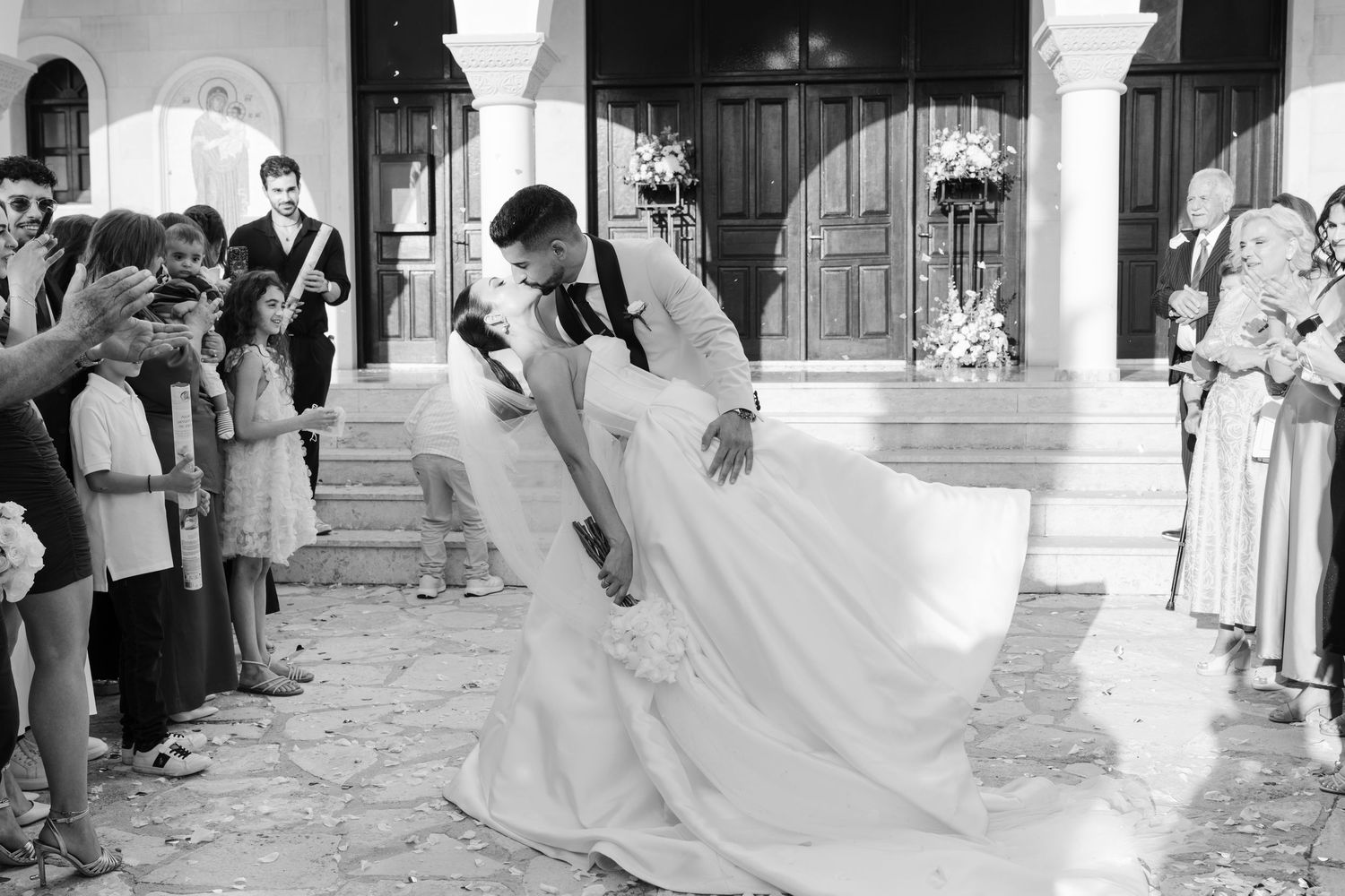 Bride and groom share a romantic dip kiss outside a church entrance surrounded by wedding guests.
