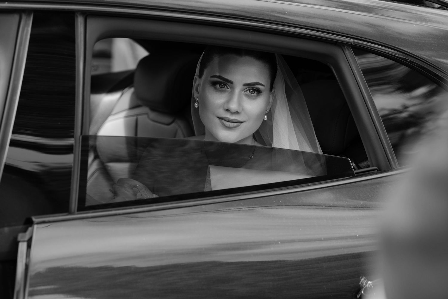 Elegant bride in veil smiling through car window on wedding day, black and white photo.