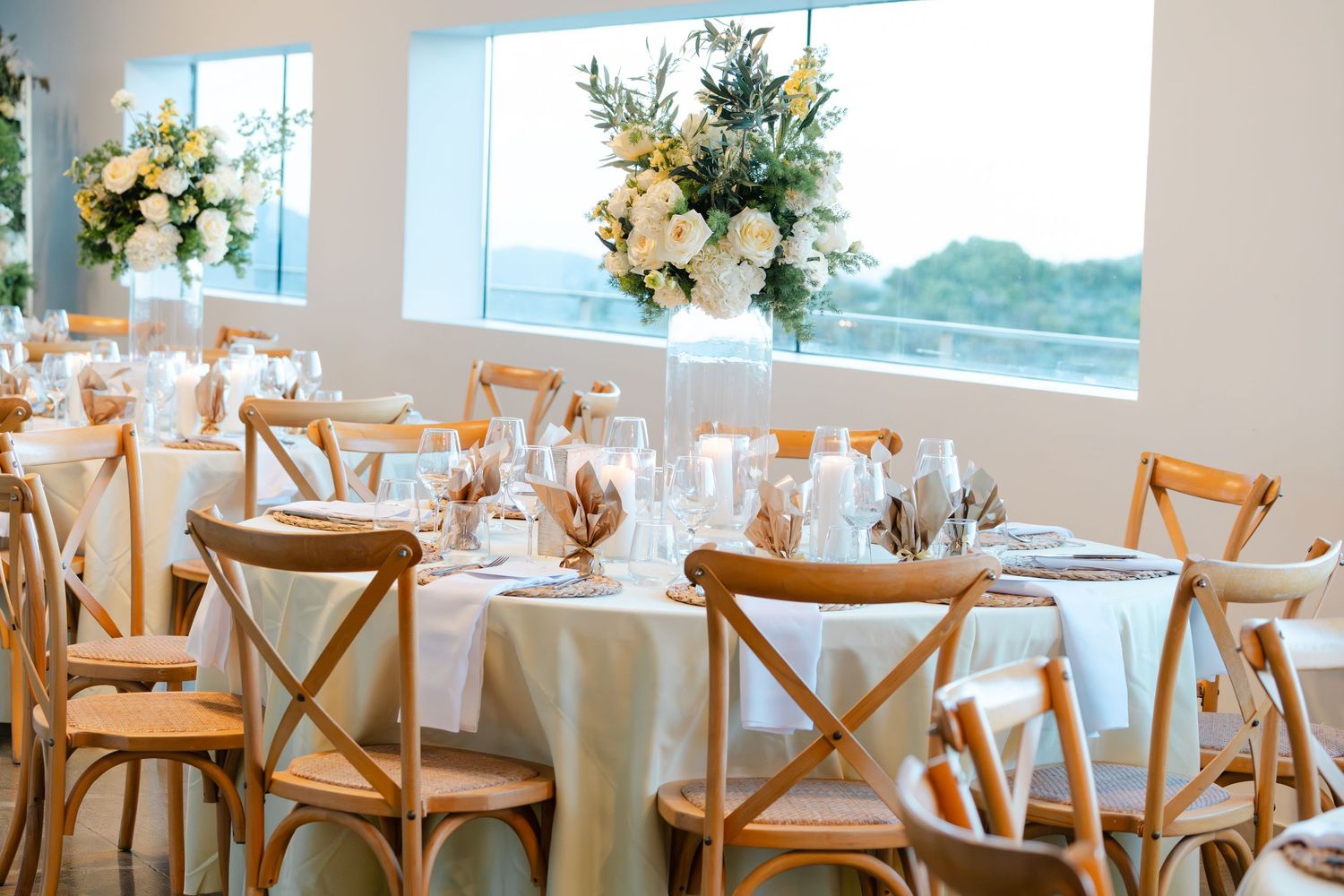 Elegant wedding reception with wooden cross-back chairs, white linen tables, and floral centerpieces by a bright window.
