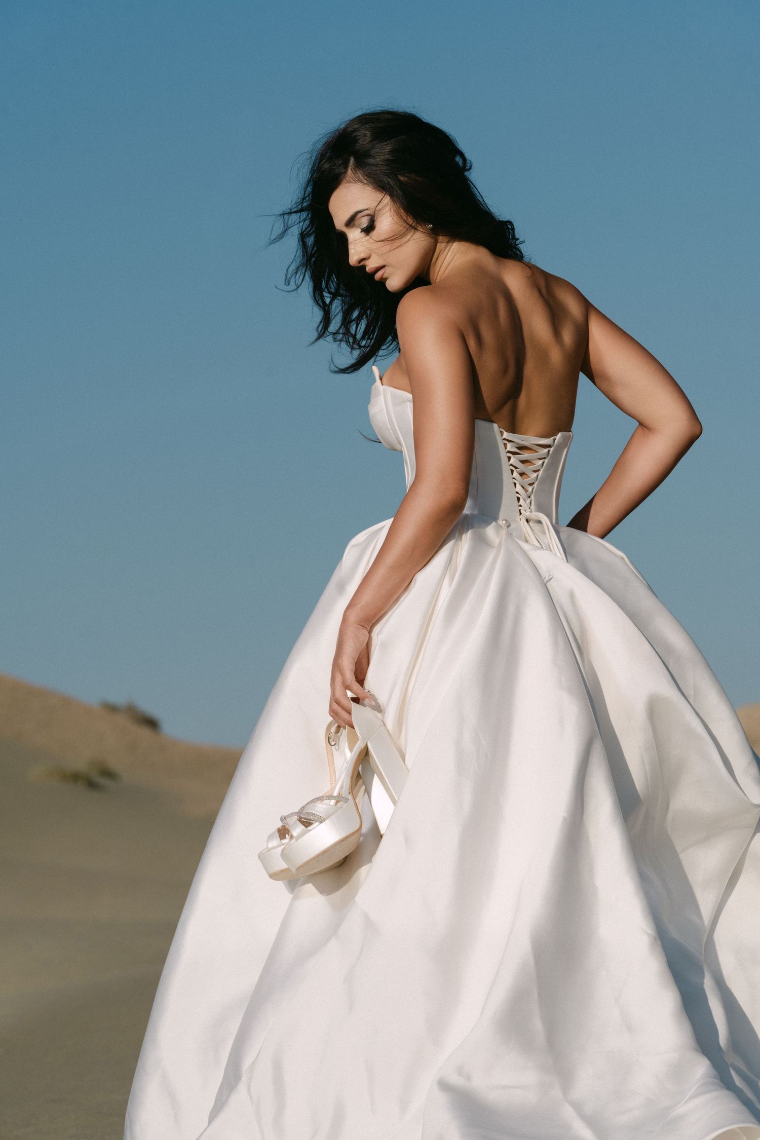 Elegant bride in strapless white ballgown holds heels while posing on desert sand dunes under blue sky.