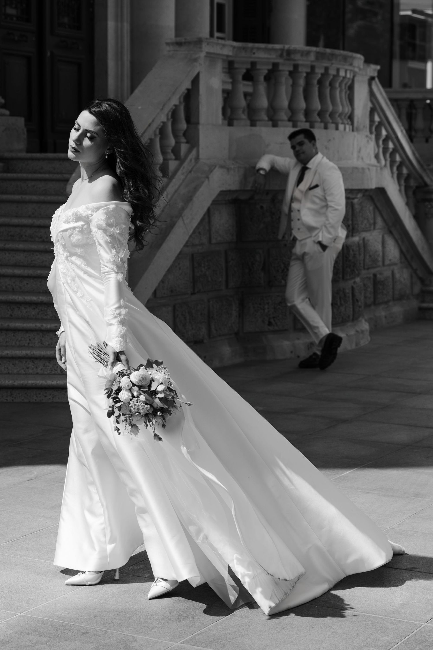 Black and white photo of bride holding bouquet with groom standing on steps in background.