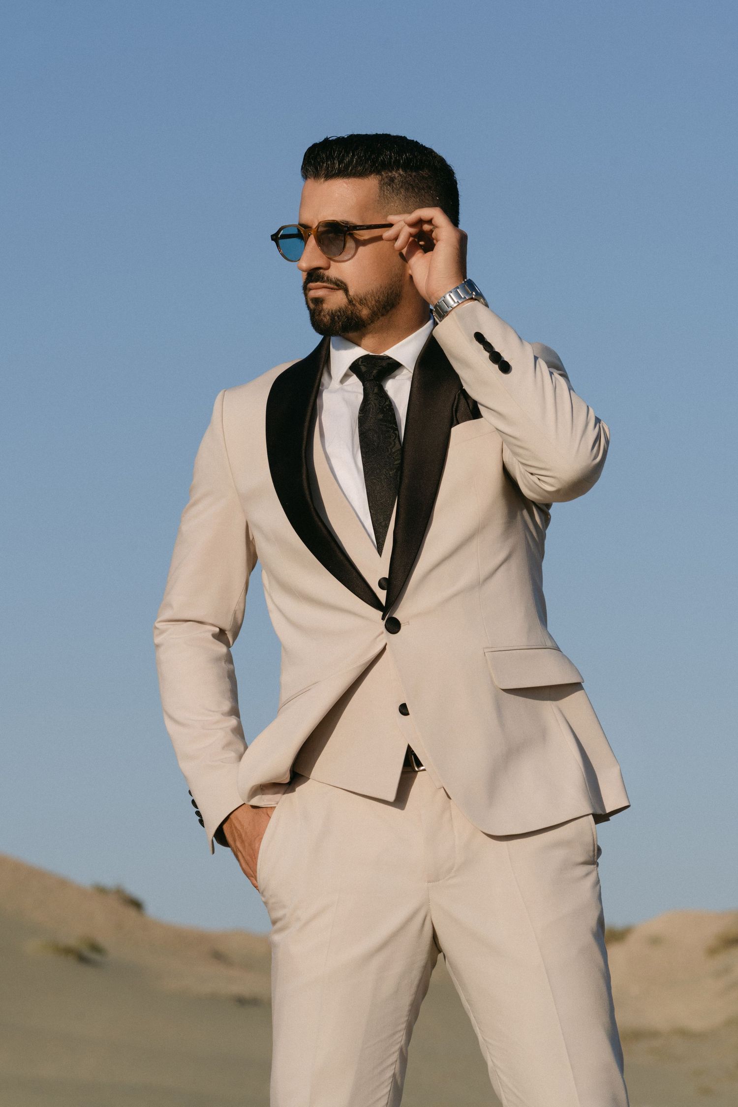 Handsome groom in beige tuxedo adjusts sunglasses while posing confidently in desert landscape.