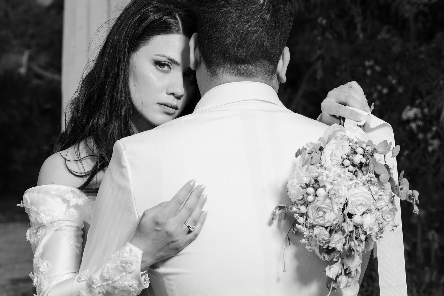 Black and white photo of bride embracing groom from behind while holding a floral bouquet.