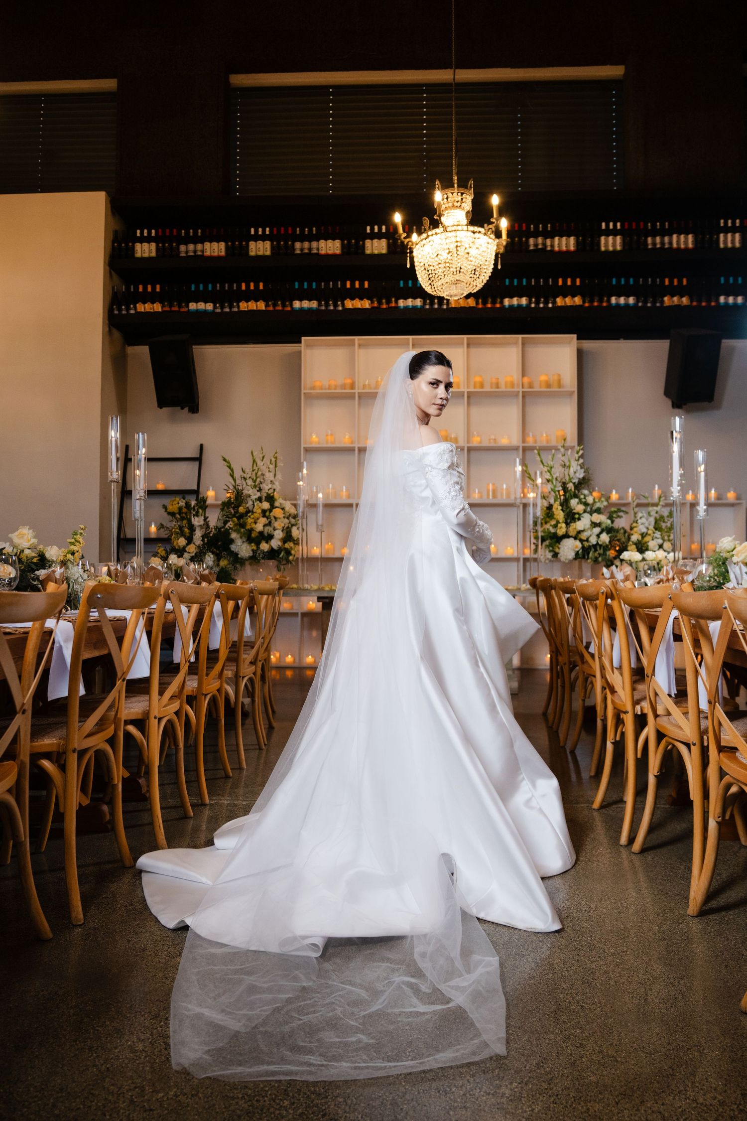 Elegant bride in white satin gown with long veil poses between decorated reception tables in candlelit venue.