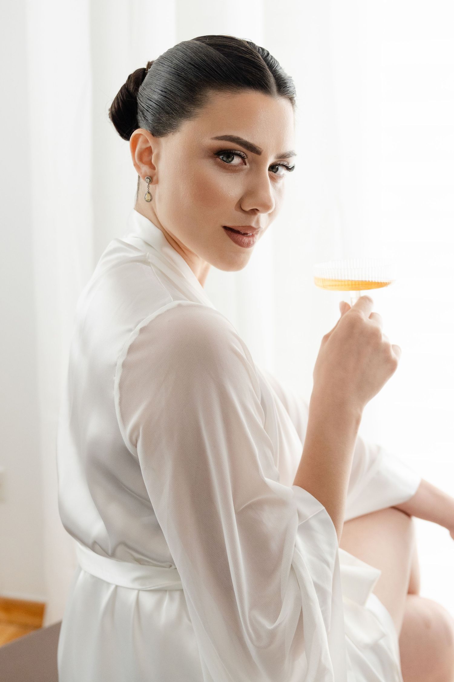 Beautiful bride in white silk robe holding champagne coupe glass before wedding ceremony.