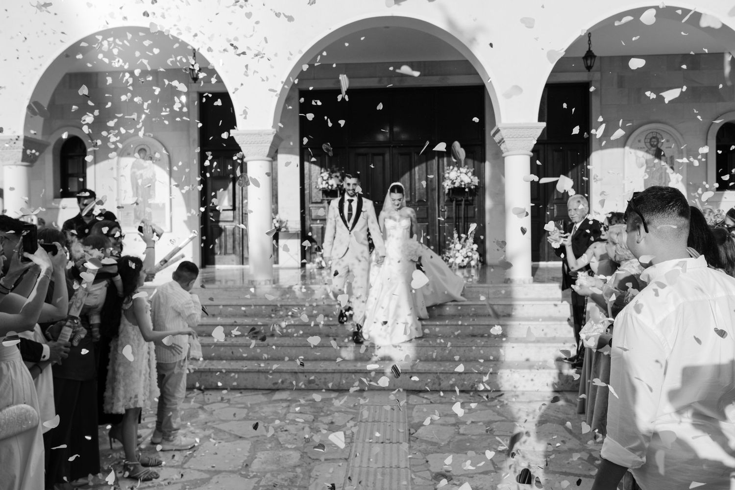 Black and white photo of newlyweds exiting church amid confetti shower from guests.