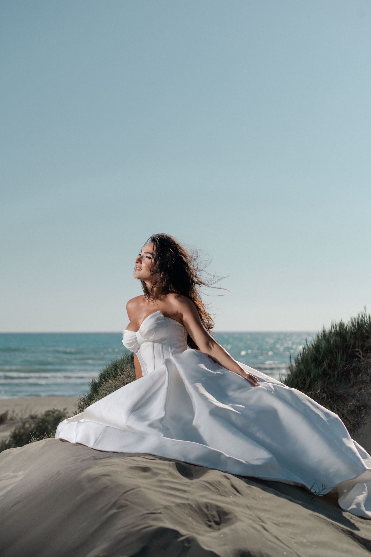 Bride in a flowing white strapless gown sits on a sandy dune with the ocean and blue sky behind her.