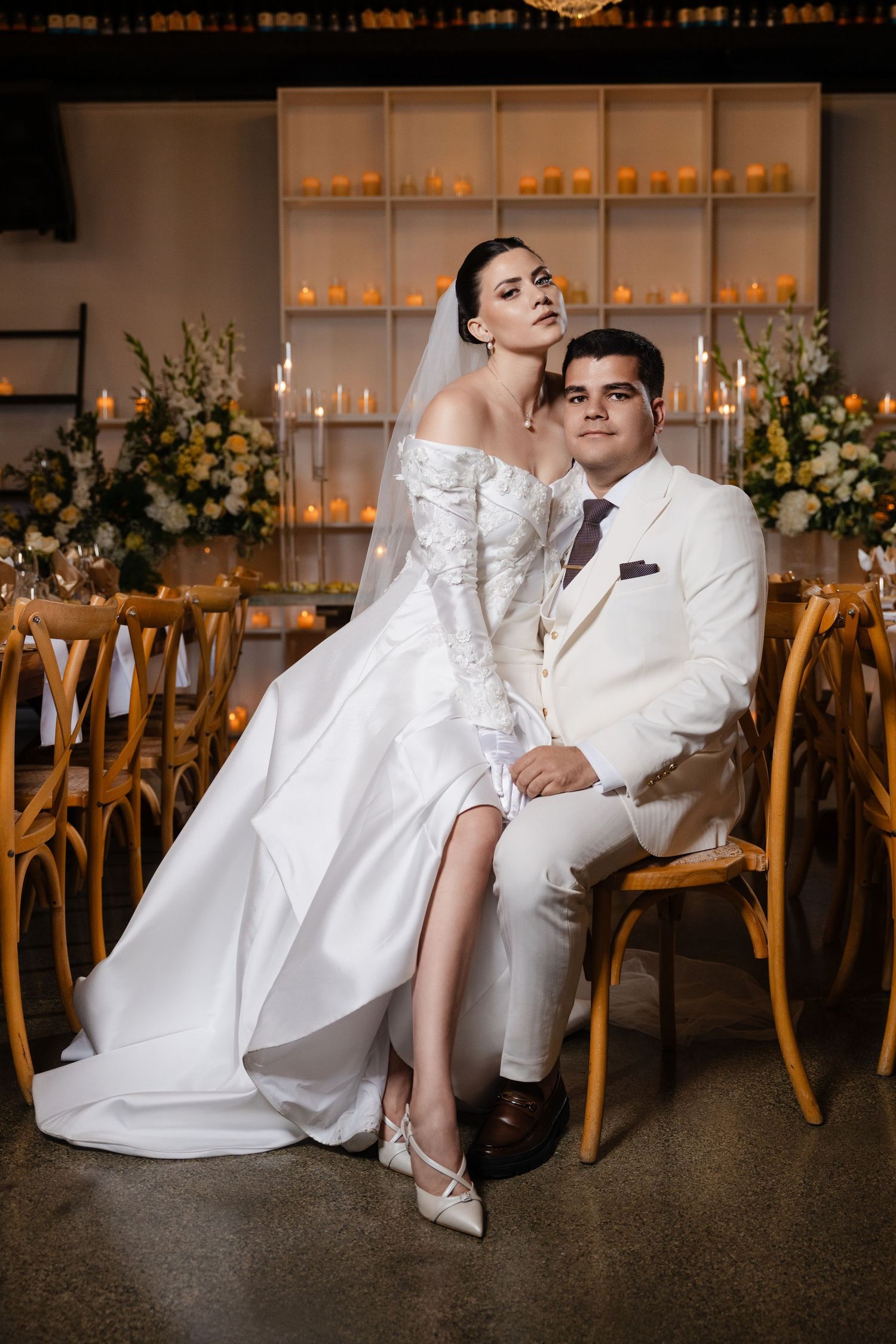 Bride and groom pose together in candlelit reception hall, she sits on his lap in white lace gown and veil.