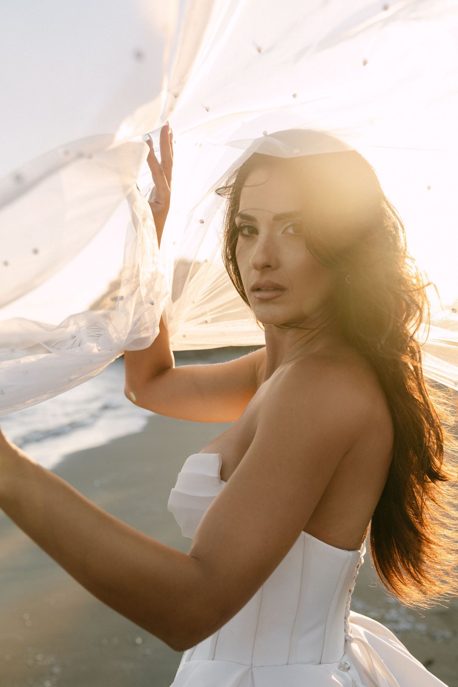 Elegant bride in white corset gown holds sheer veil with stunning golden backlit sunlight.