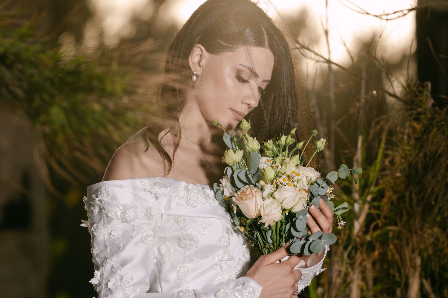 Beautiful bride with dark hair smells delicate pastel bouquet of roses and eucalyptus in soft light.