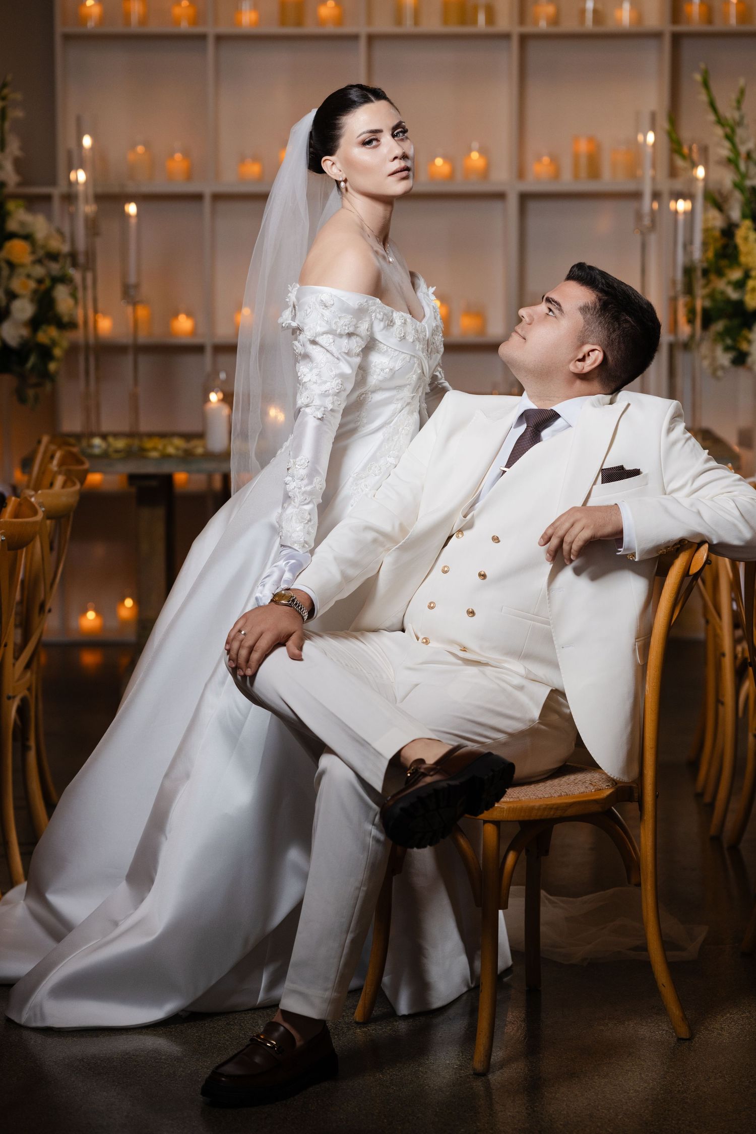 Bride in elegant white gown and groom in white suit pose dramatically in a warmly lit wedding venue.
