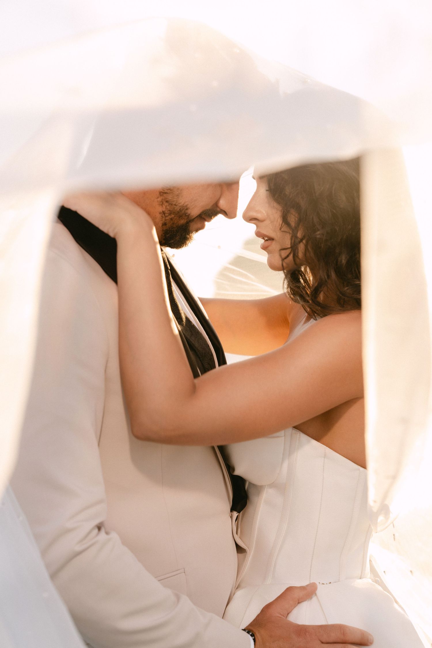 Romantic bride and groom nearly kissing framed by billowing white veil in soft golden light.