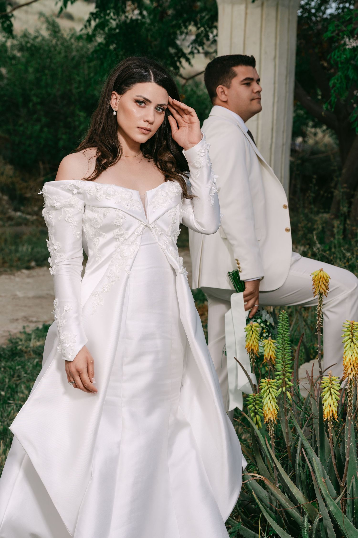 Bride in off-shoulder white gown with floral details poses with groom in white suit near garden columns.