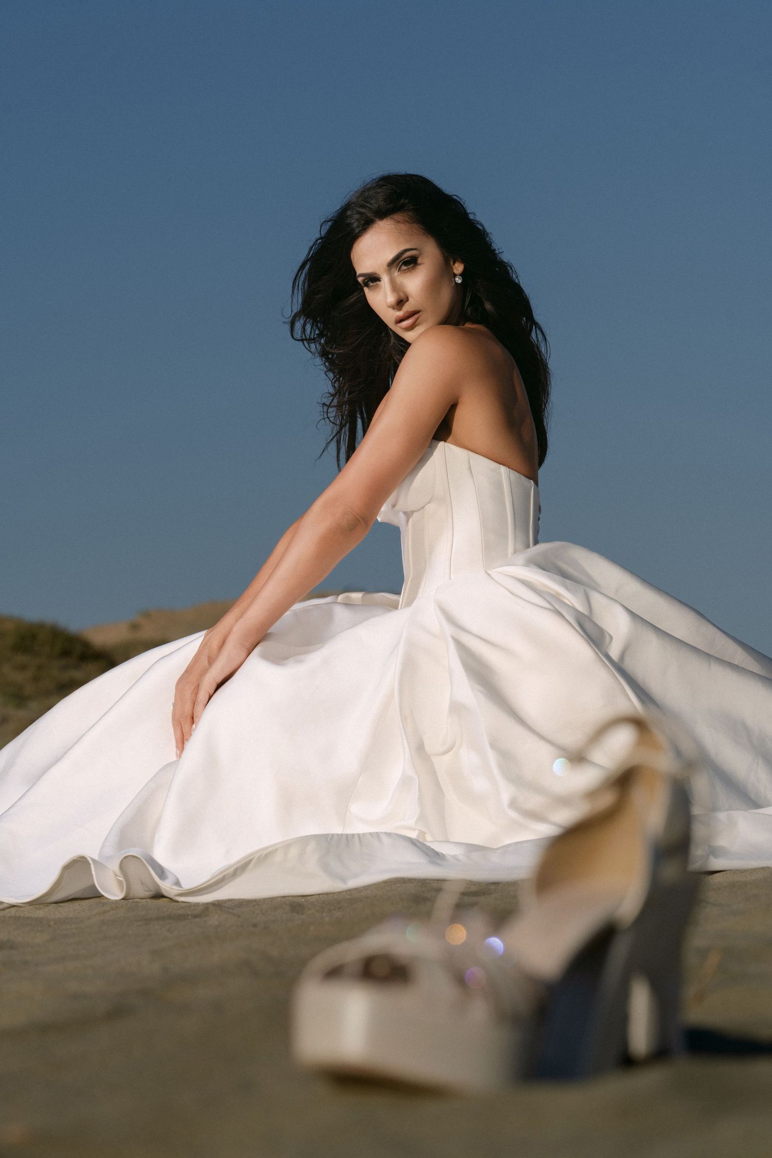 Beautiful dark-haired bride in white strapless gown sits on desert sand with platform heels in foreground.