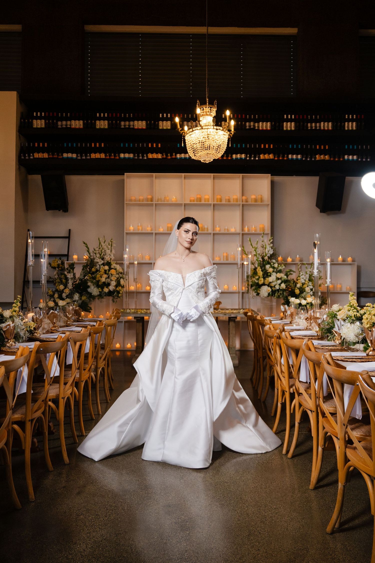 Bride in off-shoulder white gown with veil stands between candlelit reception tables in an elegant venue.