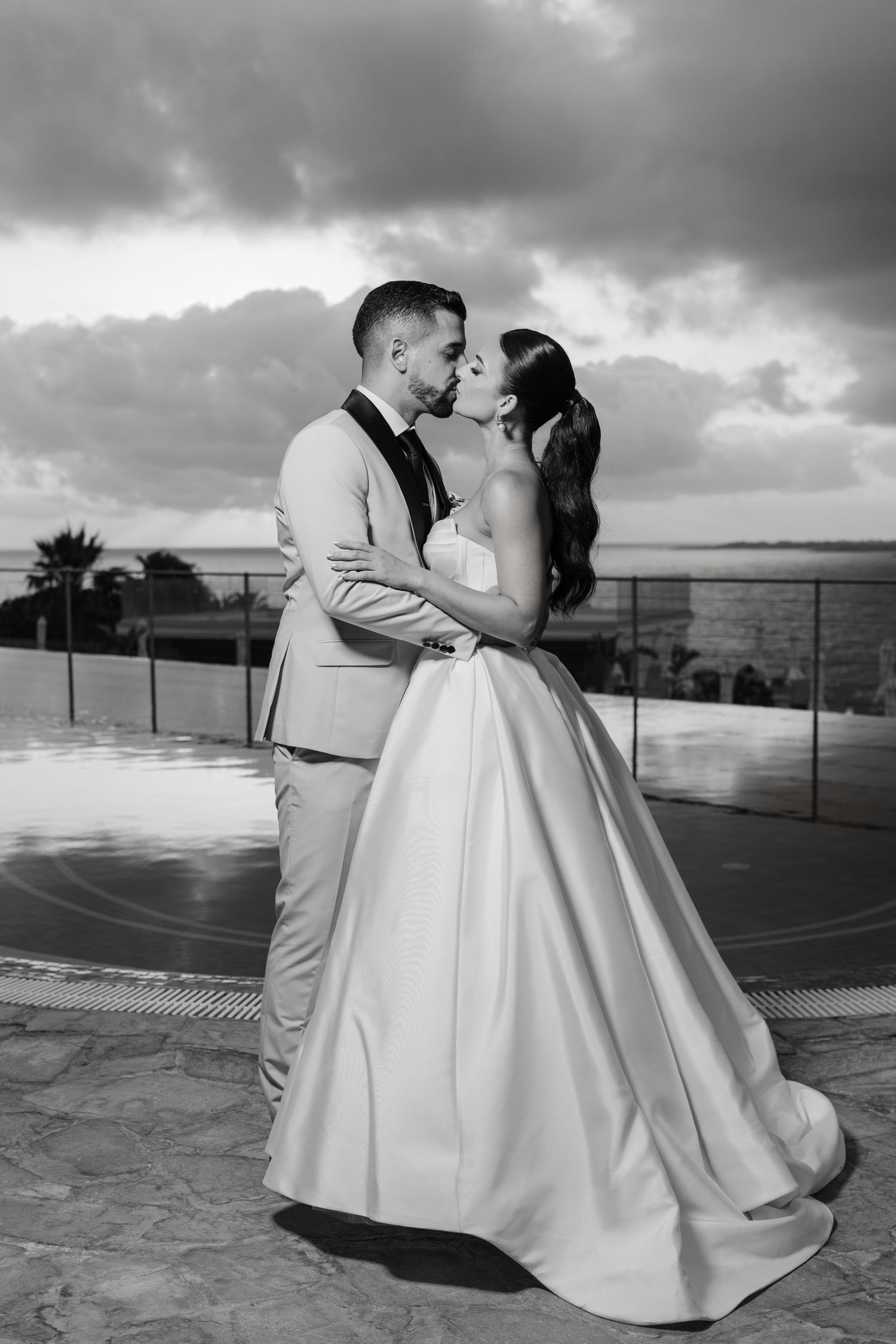 Bride and groom embrace and kiss by an infinity pool under dramatic cloudy skies.
