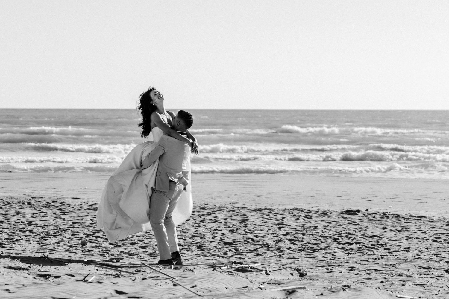 Black and white photo of groom lifting bride in a wedding dress on a beach with ocean waves.