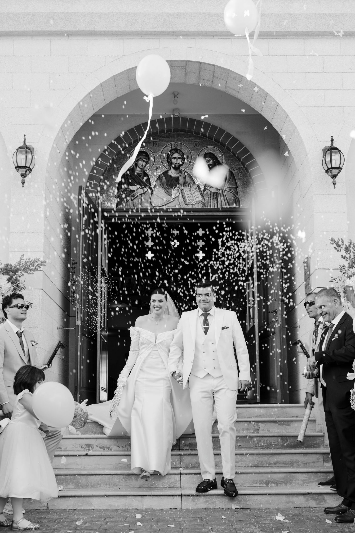 Newlyweds exit church doors as guests celebrate with confetti and white balloons.