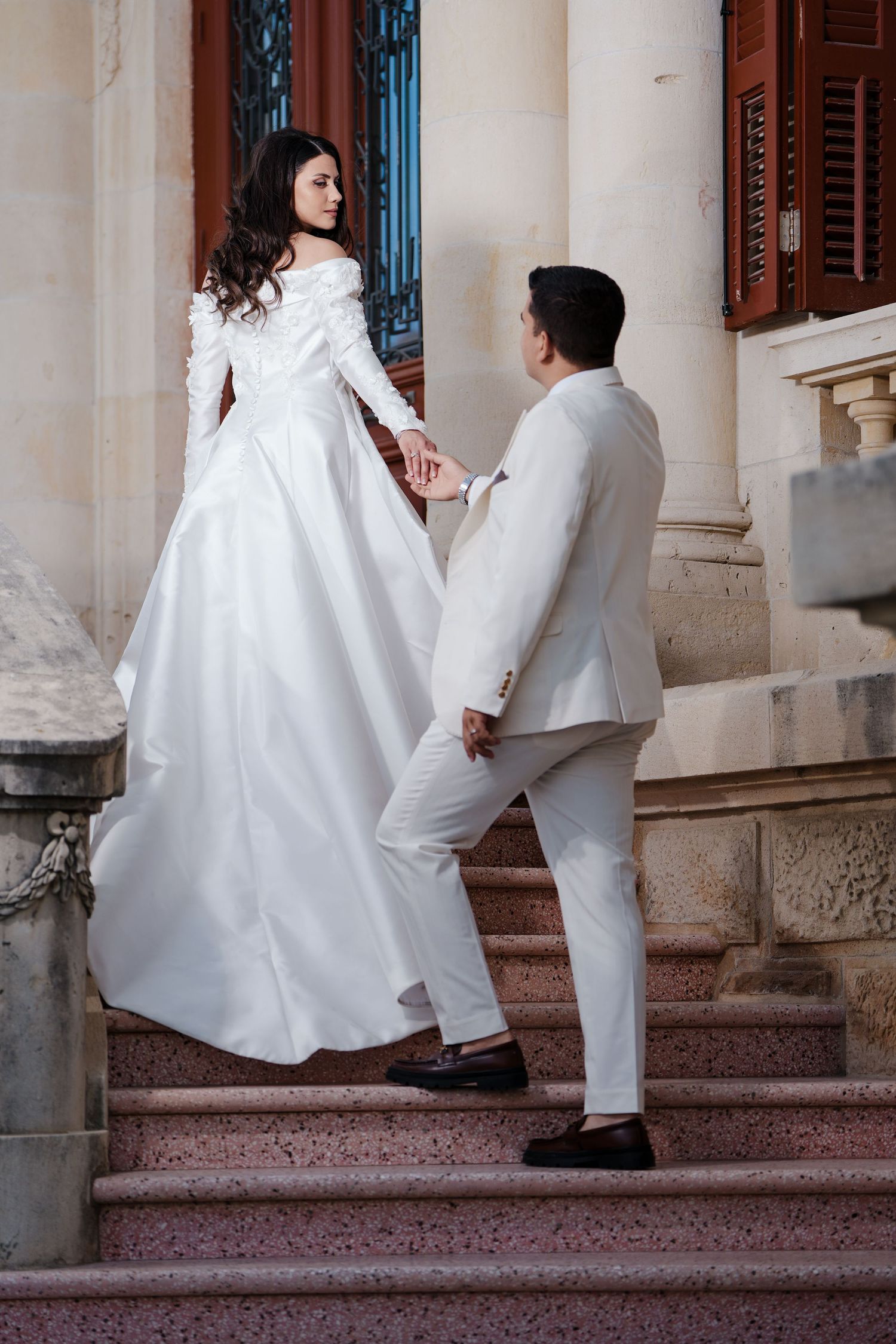Bride and groom holding hands on grand stone steps of a classical architectural building.