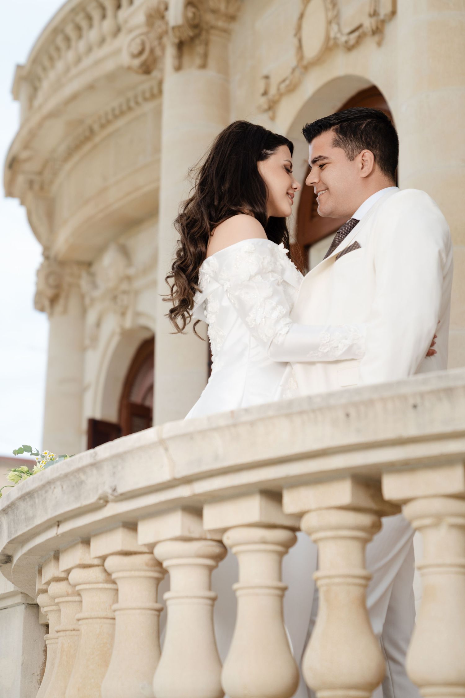 Happy bride and groom embracing and smiling at each other on an ornate stone balcony.