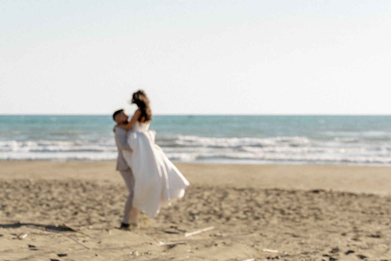 Groom lifts smiling bride in a white dress on a sandy beach with calm ocean waves behind them.