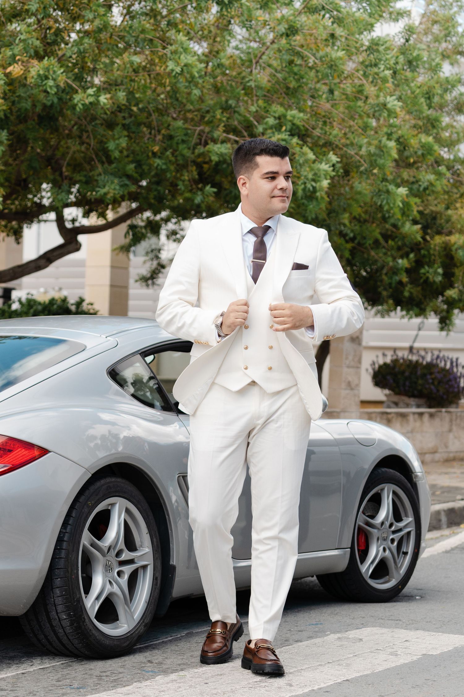 Stylish groom in white three-piece suit leaning against silver sports car outdoors.