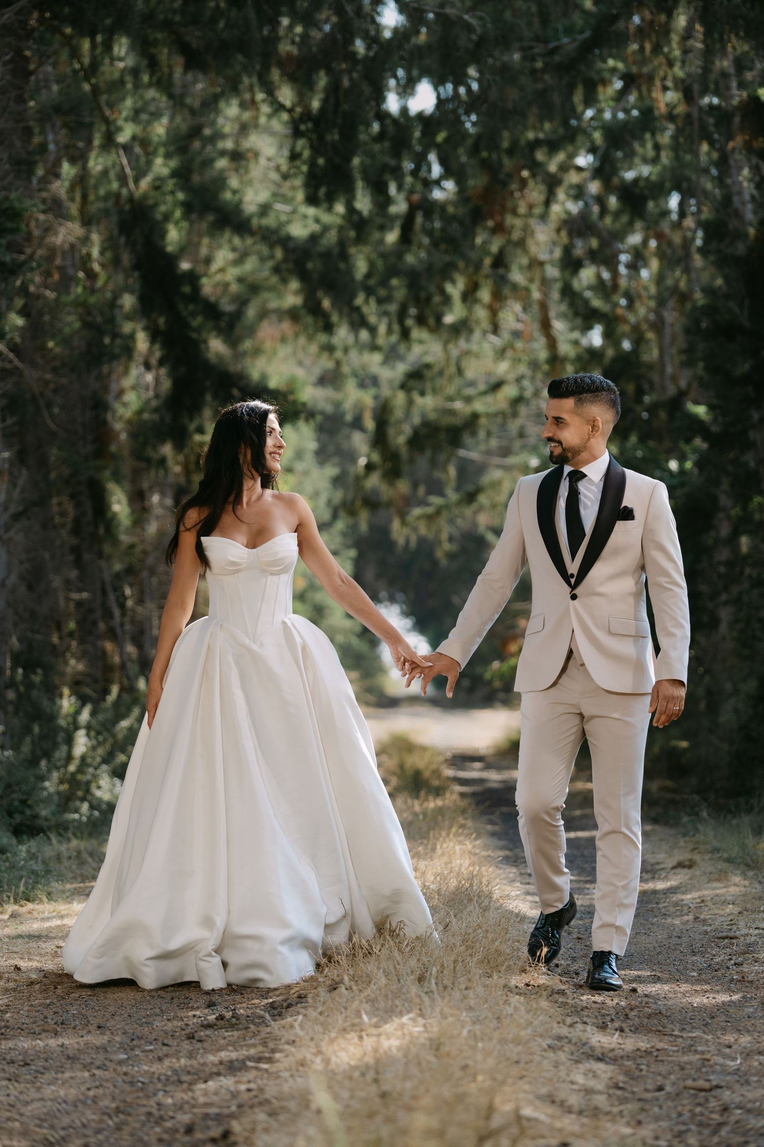 Bride and groom hold hands walking on a forest path, she in a white ballgown, he in a cream tuxedo.